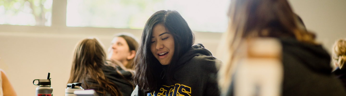 a student in a Regis University hoodie smiles while participating in a classroom discussion, surrounded by peers