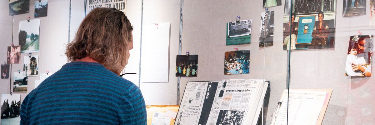 Person in a striped blue shirt on the left looks into a glass case of books, papers and photos