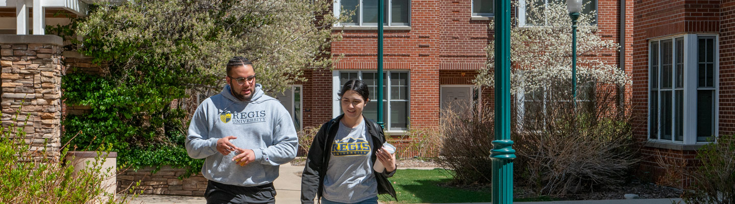 two students, engaged in conversation, walk down a courtyard sidewalk outside of a brick student housing facility