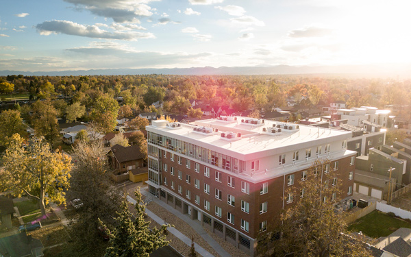 Aerial view of a modern brick and glass residential building surrounded by trees and a suburban neighborhood at sunset.