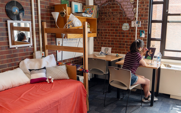 a student paints at a desk in a residence hall room with bunk beds, brick walls, and large windows letting in natural light