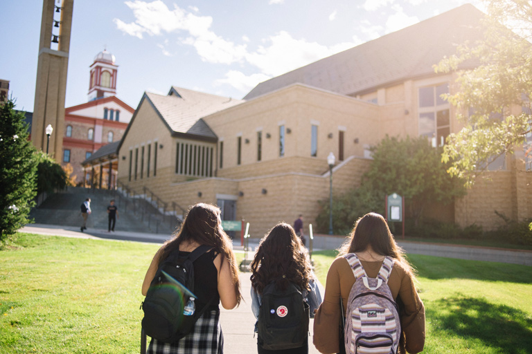 three students with backpacks walk toward a sunlit academic building at Regis University, with the campus bell tower in the background