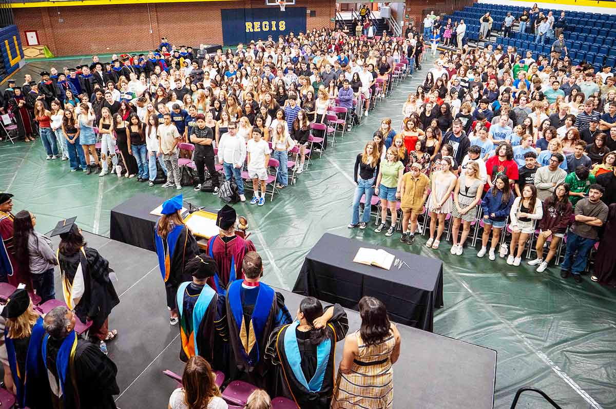 a large group of diverse students, including international students, stands on the court of the Regis Field House while enjoying Fall Convocation ceremonies