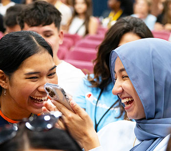 one student holds up a phone as she and her friend laugh during Convocation in the Regis Field House