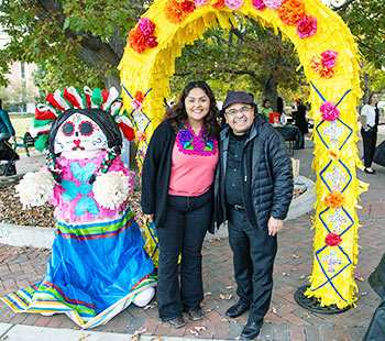 a student and faculty member pose beneath colorful Dia de Los Muertos decorations on the Regis campus