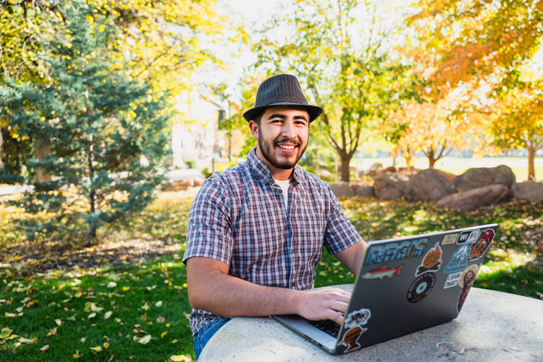 a smiling student in a plaid shirt and fedora works on a laptop covered in stickers while sitting outdoors on the Regis University campus