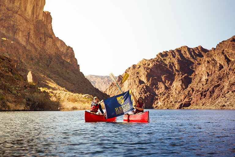 Two students in canoes on a lake hold up a Regis flag during an outdoor experience trip in beautiful Colorado