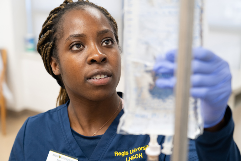 nursing student in a navy scrubs uniform inspects an IV bag, wearing gloves and a name badge for Regis University LHSON