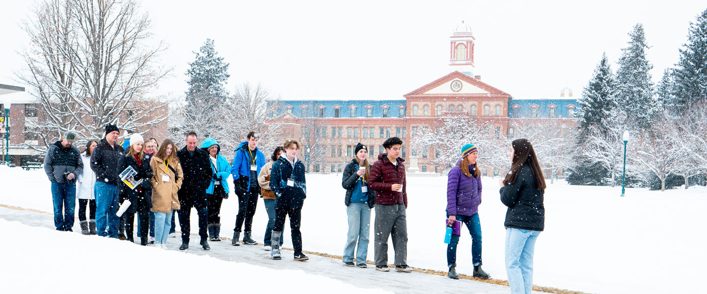a campus tour group walks through the snow at Regis University listening to a guide with Main Hall in the background