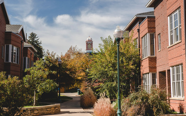 Brick residence hall buildings line a tree-filled walkway, with a bell tower visible in the distance.