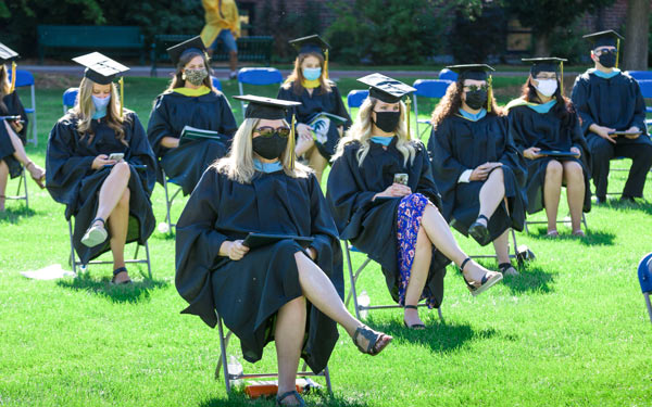 Graduates seated outdoors in socially distanced chairs wearing caps, gowns, and face masks during Regis University's 2020 commencement ceremony.
