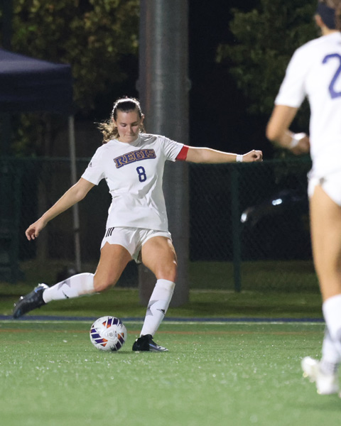 Cullinan in a white uniform with the number 8 and the word "Regis" on the front, preparing to kick a soccer ball during a night game. She wear a red captain's armband and black cleats, with another player partially visible in the foreground.