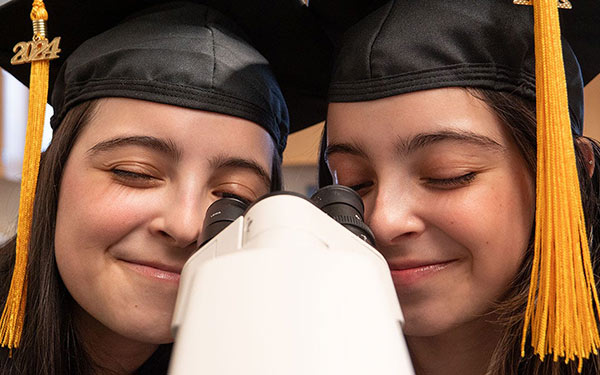 Isabella and Katelyn De Leon look through a microscope while wearing their 2024 graduation caps