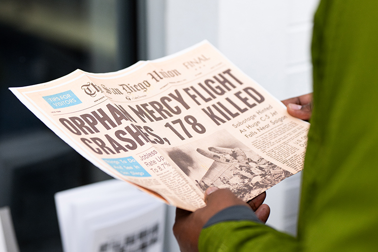 an exhibit attendee holds an archival newspaper with the headline "Orphan Mercy Flight Crashes, 178 Killed" at the Operation Babylift exhibit in the Dayton Memorial Library