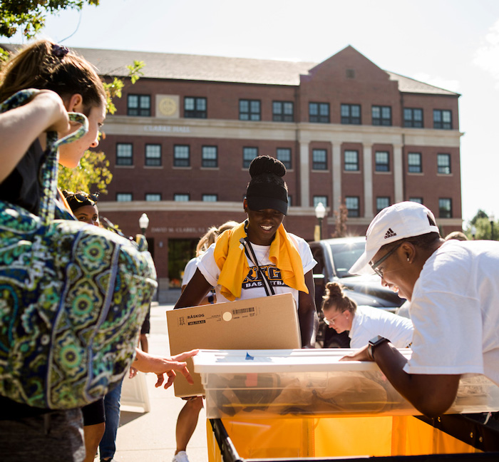 Students help each other move into a residence hall on a sunny day, unloading boxes and bins with smiles.