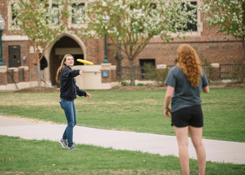 Two students play frisbee on a green campus lawn, with blossoming trees and a brick building in the background.