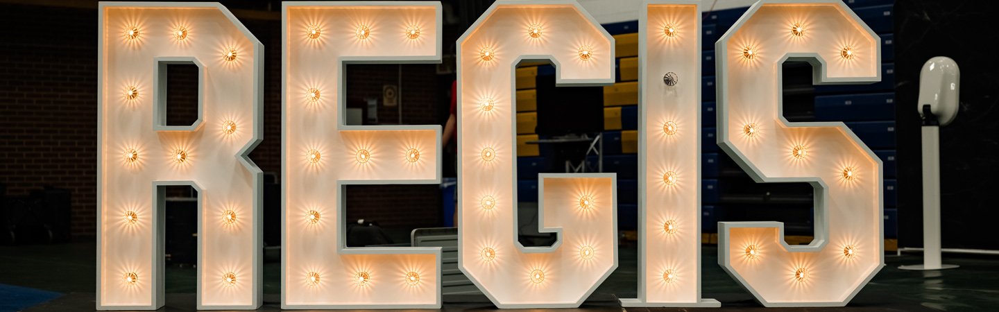 Large illuminated marquee letters spelling REGIS displayed indoors during Blue and Gold Weekend at Regis University.