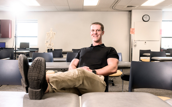A student in a black polo shirt leans back with feet up in a classroom with tables and chairs.
