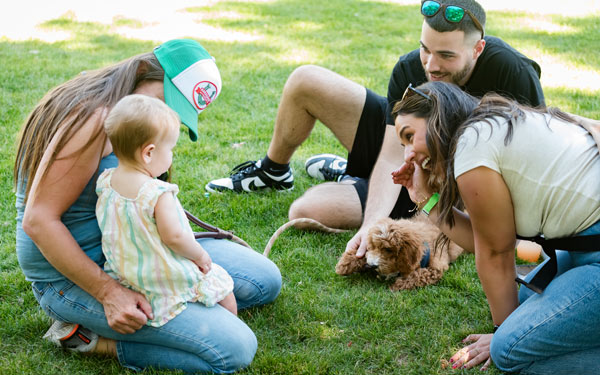 A group of people sitting on the grass during a sunny day at the Blue and Gold event. A baby in a striped dress is interacting with a fluffy brown puppy, while three adults—two women and a man—watch and smile. The setting is casual and friendly, with everyone enjoying the moment outdoors.