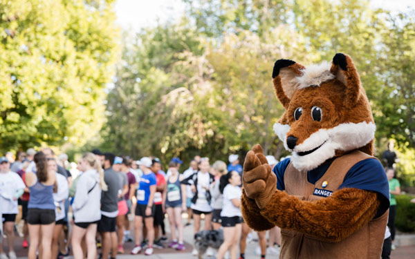 egi the fox mascot claps and smiles in front of a crowd of runners preparing for a race during the Blue and Gold event. The outdoor scene is lively and festive, with participants in athletic wear gathered under sunny skies and surrounded by green trees.