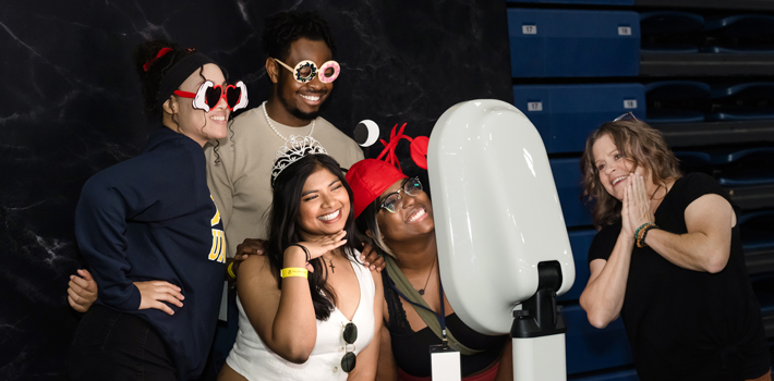 A group of smiling students and an adult pose with playful props and accessories in front of a photo booth during a Blue and Gold Weekend event at Regis University.