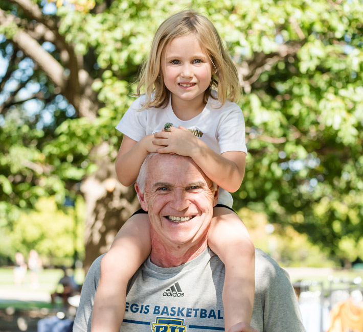 A smiling older man wearing a Regis Basketball shirt carries a young girl on his shoulders at a sunny outdoor event on the Regis University campus during Blue and Gold Weekend.