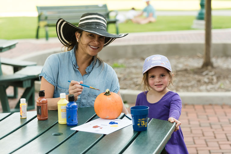 A woman and a young girl smiling while painting a pumpkin at an outdoor table during Blue and Gold Weekend at Regis University. Paint bottles, a paintbrush, and a Regis-themed cup are on the table.