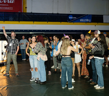 A crowd of students and guests gather in a gymnasium, chatting and enjoying the festivities during Regis University's Blue and Gold Weekend.