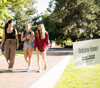 Three young women walk and laugh along a sunny brick pathway on the Regis University campus, passing a 'Welcome Home! Blue & Gold Weekend' sign.