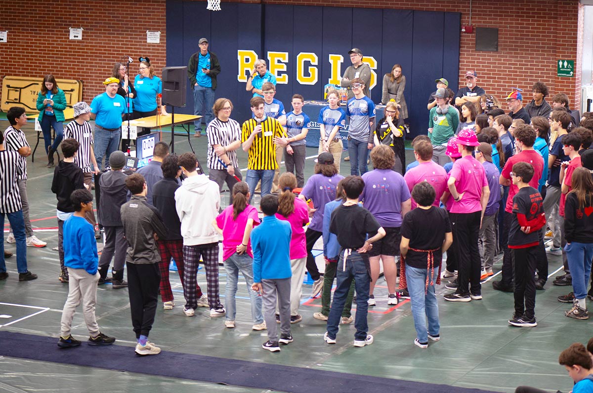 a large crowd stands on the floor and sits in the stands of in the Regis Field House as competitors in the First Tech Challenge hold microphones and present their projects.